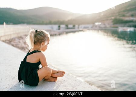 Das kleine barfuß Mädchen sitzt im Kreuz auf dem Pier und schaut aufs Wasser. Seitenansicht Stockfoto