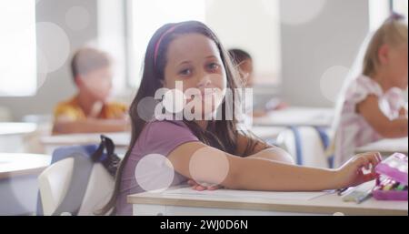 Bild von Bokeh-Lichtflecken über glücklichen birassischen Schulmädchen, die in der Klasse am Schreibtisch sitzen Stockfoto