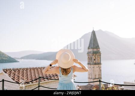 Das Mädchen steht auf der Aussichtsplattform in der Nähe des Glockenturms und hält ihren Hut mit den Händen. Perast, Montenegro. Rückansicht Stockfoto