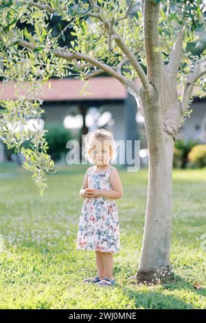 Das kleine Mädchen steht neben einem jungen Baum im Garten mit gefalteten Händen vor ihr Stockfoto