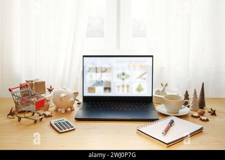 Online-Shopping im Urlaub, Laptop auf einem Holzschreibtisch mit Weihnachtsdekoration, Schweinebank, Taschenrechner und Notizblock, um Geschenke zu finden Stockfoto