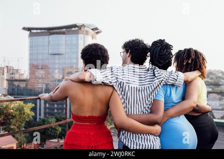 Vier Freunde umarmen sich und stehen mit dem Rücken auf dem Dach des Hauses. Mann und afrikanische Frauen in lässiger Kleidung schauen sich die Stadt an Stockfoto