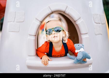 Ein kleines Mädchen in einer Sonnenbrille mit einem Spielzeug in der Hand schaut aus dem Fenster einer Spielzeugburg auf dem Spielplatz Stockfoto