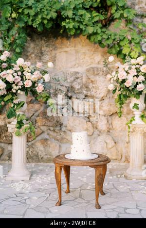 Hochzeitstorte steht auf einem Holztisch in der Nähe eines Hochzeitshalbbogens in der Nähe einer Steinmauer im Garten Stockfoto