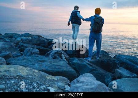 Ein junges Paar spaziert bei Sonnenuntergang am Meer entlang. Ein Mann und eine Frau in lässiger Kleidung stehen zurück. Stockfoto