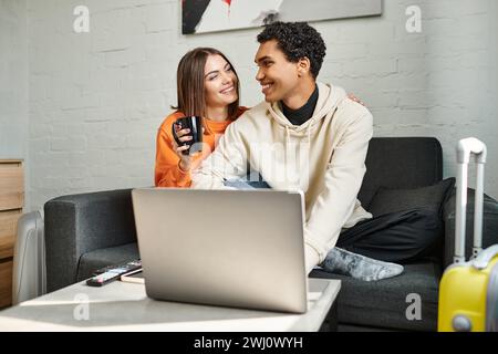 Sorglose interrassische Paare teilen einen gemütlichen Moment, während sie auf der Couch sitzen und Laptop, Hostel benutzen Stockfoto