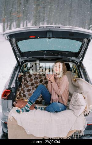Die junge Frau sitzt mit einer Tasse Kaffee, die vor Lust im Kofferraum eines Autos in einem Winterwald schwingt Stockfoto
