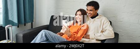 Glückliche, vielfältige Paare teilen einen gemütlichen Moment, indem sie auf einer bequemen Couch durch die Telefone scrollen, Banner Stockfoto