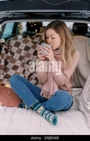 Das Mädchen sitzt im Kreuz auf einer Decke im Kofferraum eines Autos und trinkt Kaffee aus einer Tasse Stockfoto