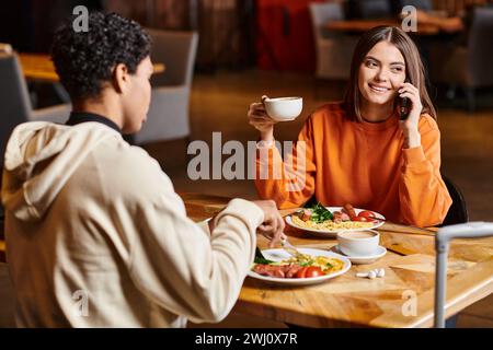 Entspannte Frau, die einen angenehmen Anruf hatte, während sie eine Tasse Kaffee in der Nähe eines schwarzen Freundes im Café hielt Stockfoto