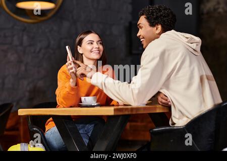 Glückliche Frau im orangefarbenen Pullover zeigt dem lächelnden schwarzen Freund das Telefon, während er im Café sitzt Stockfoto