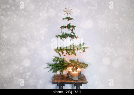 Christbaumalternative, immergrüne Zweige mit Dekoration, die an der Wand über einem rustikalen Holzhocker mit Kerzen hängen Stockfoto