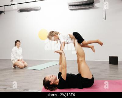 Budva, Montenegro - 5. august 2023: Trainer hebt ein kleines Mädchen auf ausgestreckten Beinen und Armen auf, während es im Fitnessstudio auf einer Matte liegt Stockfoto