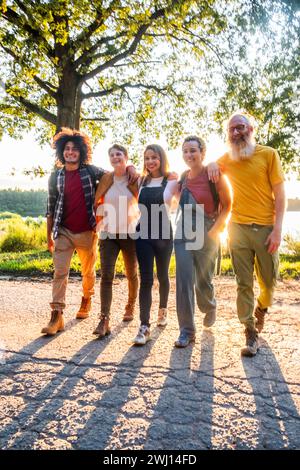 Dieses einladende Bild zeigt eine Familie mehrerer Generationen, die in den späten Stunden eines sonnigen Tages einen gemütlichen Spaziergang zusammen macht. Die Familie ist unterschiedlich im Alter, von einem jungen Afroamerikaner mit lockigen Haaren bis zu einem älteren Herrn mit weißem Bart und gelbem Hemd. Sie sind lässig gekleidet, was auf einen entspannten Ausflug hinweist. Das warme Sonnenlicht wirft lange Schatten auf den Weg hinter ihnen und beleuchtet ihre Gesichter mit einem sanften Leuchten, was das Gefühl einer gemütlichen Familienaktivität am Ende des Tages verstärkt. Der große Baum im Hintergrund und der offene Himmel deuten darauf hin, dass sie sich in einem Park oder einem natürlichen Gebiet befinden, das förderlich ist Stockfoto