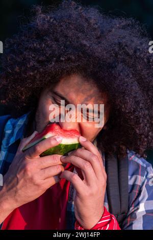 Das Bild zeigt einen jungen Nordafrikaner, Tunesier mit einem dichten, lockigen Afro-Haar, der einen Biss einer Reifen Wassermelonenscheibe tief verkostet. Sein Gesichtsausdruck ist ein Ausdruck reiner Freude und Zufriedenheit, während er in die Wassermelone beißt, mit fest geschlossenen Augen und geknitterten Augenbrauen, was die Intensität des Geschmackserlebnisses unterstreicht. Er trägt ein rotes T-Shirt mit einem blau-weiß karierten Hemd, das nicht geknöpft ist, um die Wärme zu bewahren. Ein schwarzer Rucksack ist sichtbar, was darauf hindeutet, dass er eine Pause während eines Ausflugs im Freien genießt. Die mittelbraune Haut des Mannes wird gegen das dunkle t gestellt Stockfoto