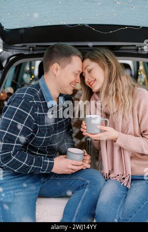 Lächelnder Mann und Frau, die die Stirn berühren, während sie mit Bechern im Kofferraum sitzen Stockfoto