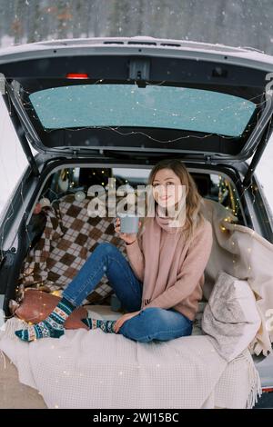 Ein lächelndes Mädchen mit einer Tasse Tee sitzt auf einer Decke im Kofferraum eines Autos unter Schneefall im Wald Stockfoto