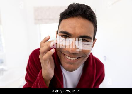 Glücklicher birassischer Mann, der Euteraugenflecken im Badezimmer zu Hause aufträgt Stockfoto