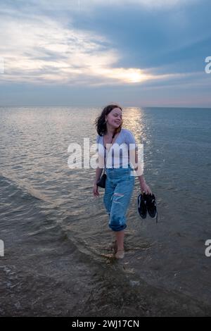 Das Bild fängt eine junge Frau ein, die bei Sonnenuntergang im ruhigen Wasser gemütlich waten kann. Mit den Schuhen in der Hand scheint sie das Gefühl des Wassers auf ihren Füßen zu genießen, während ihr Blick auf das warme Licht der absteigenden Sonne gerichtet ist. Ihr entspanntes Auftreten und das lässige Outfit deuten auf einen spontanen Moment der Verbindung mit der Natur hin. Der Himmel, eine Leinwand aus sanftem Blau und Violett mit den goldenen Farbtönen der Sonne am Horizont, bietet eine malerische Kulisse für diese friedliche Szene. Eine junge Frau, die bei Sonnenuntergang im Wasser watet. Hochwertige Fotos Stockfoto