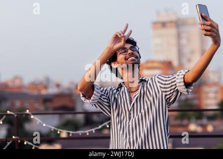 Junge Lockenhaarige in Brille und gestreiftem Hemd nimmt Videoanrufe am Telefon an. Ein lächelnder Mischlingsmann zeigt das Siegessymbol und Stockfoto
