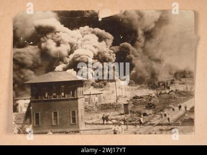 TULSA RACE RIOT (31. Mai und 1. Juni 1921): In Tulsa, Oklahoma, griffen Mobs weißer Einwohner brutal die afroamerikanische Gemeinschaft an, um Ein Schwarz-weiß-Foto des Greenwood District in Tulsa, Oklahoma, das während des Tulsa Race Massakers 1921 verbrannt wurde. Das Bild zeigt ein Gebäude und mehrere Personen im Vordergrund, die große Wolken von dunklem Rauch im Hintergrund beobachten. Das Foto ist an den Ecken gebogen und hat an der oberen Mittelkante einen Verlust Stockfoto