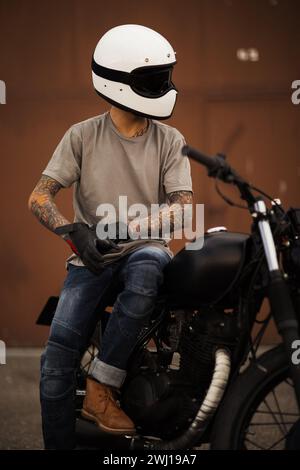 Ein getarnter Radfahrer im Helm sitzt auf einem maßgeschneiderten Motorrad im urbanen Hintergrund Stockfoto