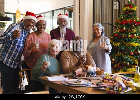 Fröhliche, vielfältige Gruppe von älteren Freunden in weihnachtsmützen, die weihnachtsessen feiern und zu Hause toasten Stockfoto