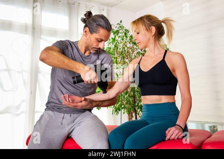 Konzentrierter Masseur in Freizeitkleidung sitzend und drückend Massagepistole auf Arm des Kunden in Aktivkleidung im Studio mit grüner Pflanze und natürlichem Licht von Stockfoto