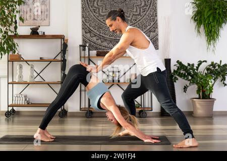 Junge fitte Frau, die Adho Mukha Svanasana Yoga übt, posiert auf Matte mit Hilfe von muskulösen männlichen Trainern, die während des Trainings im modernen Studio stehen Stockfoto