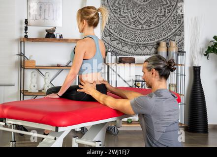 Seitenansicht einer fokussierten Physiotherapeutin mit Pferdeschwanz, die den unteren Rücken einer Patientin in Aktivkleidung untersucht, die während der Behandlung in m auf roter Bahre sitzt Stockfoto