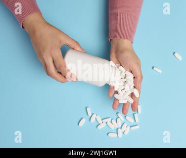 Die Hand einer Frau, die eine Plastikflasche und weiße ovale Pillen auf blauem Hintergrund hält, von oben. Stockfoto