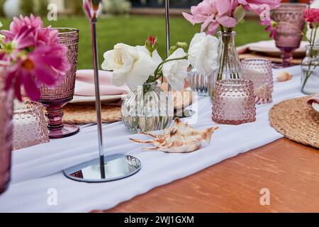 Pinkfarbene Picknicktische, Blumenarrangements, Muscheln aus nächster Nähe. Gemütliche Einrichtung mit Blumen und Kerzen. Stockfoto