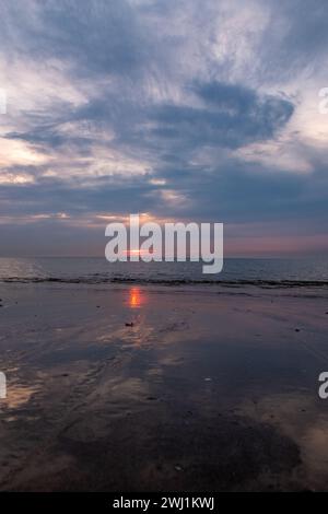 Dieses Bild fängt die stimmungsvolle Atmosphäre eines Sonnenuntergangs mit einem dramatischen Himmel ein, wo die Wolken mit blauen, grauen und dezenten warmen Tönen gemalt sind. Der letzte Teil der Sonne taucht in Richtung Horizont und reflektiert den nassen Sand des Strandes, der als natürlicher Spiegel fungiert. Das ruhige Meer in der Ferne trifft auf den Himmel und schafft eine weiche, gestreute Linie, die die ruhige und doch düstere Atmosphäre der Szene unterstreicht. Dieses Bild verkörpert die reflektierende und oft introspektive Stimmung, die ein Sonnenuntergang am Strand hervorrufen kann. Dramatischer Himmel bei Sonnenuntergang, der sich auf dem nassen Strand spiegelt. Hochwertige Fotos Stockfoto