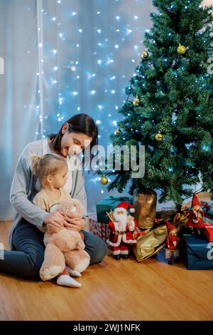 Ein kleines Mädchen mit einem Teddybären sitzt auf dem Schoß ihrer Mutter auf dem Boden und schaut sich die Geschenke unter dem Baum an Stockfoto