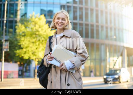 Bild einer jungen, schönen Frau, die lächelnd ist, Notizbücher und Hausaufgabenunterlagen hält, neben dem Universitätsgebäude steht, Stockfoto