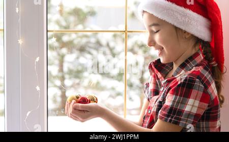 Ein Mädchen mit einem Weihnachtsmann-Hut sitzt auf der Fensterbank eines Hauses in der Nähe des Weihnachtsbaums und hält Glaskugeln in der Hand. Kind ist hav Stockfoto