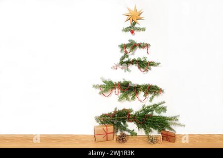 DIY-Weihnachtsbaum aus natürlichen Zweigen, die an eine weiße Wand genagelt sind, rote Dekorationskette und ein Papierstern auf der Oberseite, Geschenke auf der Stockfoto