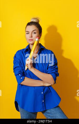 Fröhliche Frau, die blaues Hemd trägt und einen riesigen Bleistift auf gelbem Hintergrund hält Stockfoto