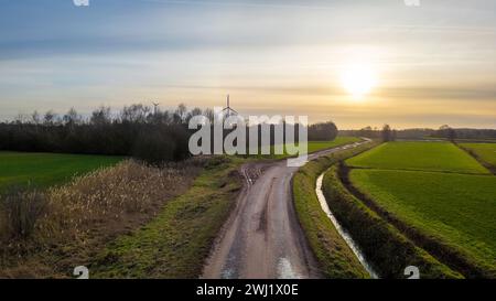 Dieses Landschaftsbild bietet einen Blick auf eine gewundene Landstraße bei Sonnenuntergang. Die Straße kurvt um ein üppiges grünes Feld und eine Reihe von Büschen, die das Auge in Richtung einer einsamen Windturbine in der Ferne führen. Die Sonne schwebt nahe am Horizont, wirft ein sanftes Licht über die Szene und schafft eine ruhige ländliche Atmosphäre. Das Vorhandensein der Windkraftanlage führt ein Element nachhaltiger Energie in die natürliche Umgebung ein und stellt einen Kontrast zwischen den traditionellen und modernen Aspekten des ländlichen Lebens dar. Sonnenuntergangshorizont Über Die Gewundene Country Road. Hochwertige Fotos Stockfoto