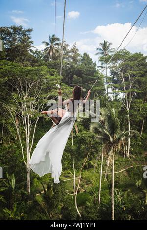 Frau in weißem Kleid, die auf Seilschaukeln schwingt, mit wunderschönem Blick auf Reisterrassen und Palmen auf der Bali Insel Stockfoto