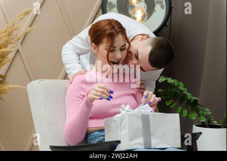 Ein Mann schenkt einem Mädchen ein Geschenk. Ein Mann umarmt ein Mädchen von hinten. Das Mädchen sitzt auf einem Stuhl. Stockfoto