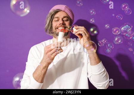 Sorgloser Mann mittleren Alters, der Seifenblasen vor violettem Hintergrund bläst Stockfoto