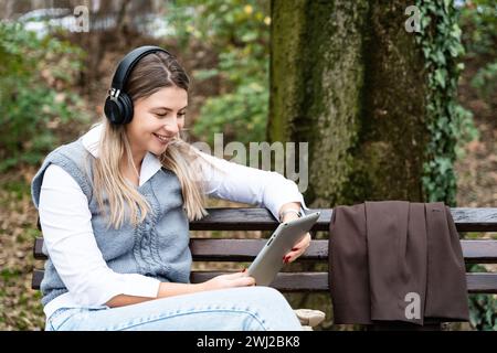 Junge Geschäftsfrau, die im Park auf der Bank sitzt und an einem digitalen Tablet arbeitet und Musik über ein schnurloses Headset hört. Geschäftsmann Freelancer macht eine Pause Stockfoto