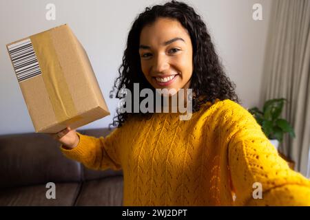 Glückliche birassische Frau in gelbem Pullover, die das Paket zu Hause hält Stockfoto