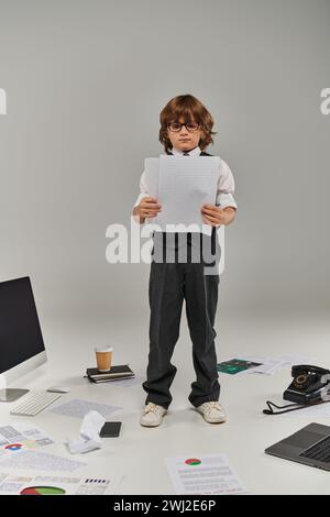 Niedliches Kind in Brille und formeller Kleidung, umgeben von Büroausstattung und Geräten, die mit Papieren stehen Stockfoto