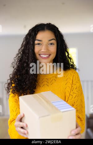 Glückliche birassische Frau in gelbem Pullover, die das Paket zu Hause hält Stockfoto