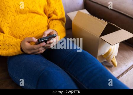 Mittelteil einer birassischen Frau in gelbem Pullover mit Paket mit Smartphone zu Hause Stockfoto