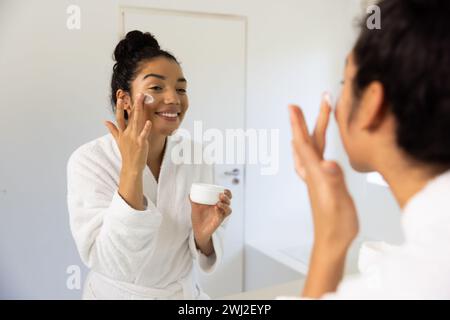 Glückliche birassische Frau im Bademantel, die im sonnigen Badezimmer Creme auf ihr Gesicht aufträgt Stockfoto