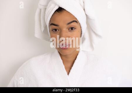 Porträt glücklicher birassischer Frau im Bademantel und mit Handtuch gegen weiße Wand Stockfoto