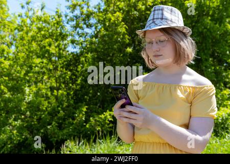 Ein asiatisches Teenager-Mädchen in gelbem Kleid, Hut und Brille kauft online am Telefon ein und kommuniziert online im Park. Ein Mädchen im Hintergrund Stockfoto